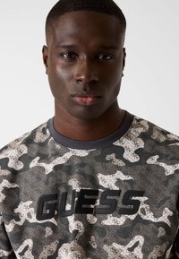 Maglietta a maniche corte con motivo mimetico, caratterizzata da tonalità di grigio e nero, con logo "GUESS" in rilievo nero sul petto.