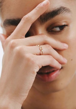 Anillo de cadena de oro con eslabones entrelazados, superficie suave, diseño delicado, llevado en una mano que cubre parcialmente la cara.