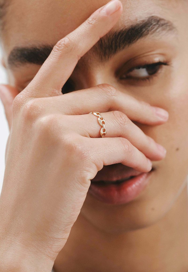Anillo de cadena de oro con eslabones entrelazados, superficie suave, diseño delicado, llevado en una mano que cubre parcialmente la cara.