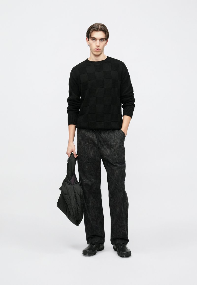 Μαύρο καρό πουλόβερ με ριμπ λαιμόκοψη, συνδυασμένο με σκούρο παντελόνι με μοτίβα. Το μοντέλο κρατάει μια μαύρη καπιτονέ ζακέτα και φοράει μαύρα παπούτσια.