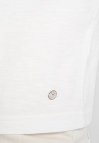 Camiseta de algodón blanca con un pequeño logo circular en el dobladillo inferior. Textura suave con un corte clásico y mangas cortas.