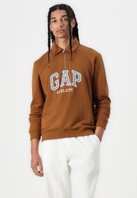 Brun zip-up hoodie med "GAP ATHLETIC" i blå bokstäver. Har en krage och ribbade mudd, tillsammans med vita byxor.