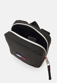 Zwarte nylon tas met een dubbele rits, voorzien van een rood, wit en blauw logo patch en een rode accent op de rits.