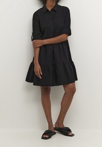 Schwarzes knielanges Kleid im Hemdblusenstil, mit aufgekrempelten Ärmeln und gestuftem Saum. Hergestellt aus weichem Stoff, mit lockerem Schnitt.