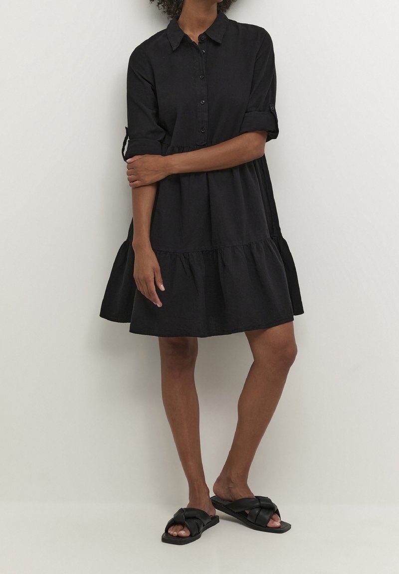 Schwarzes knielanges Kleid im Hemdblusenstil, mit aufgekrempelten Ärmeln und gestuftem Saum. Hergestellt aus weichem Stoff, mit lockerem Schnitt.