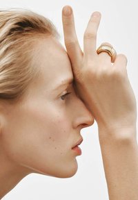 Anillo dorado estriado en una mano que toca la frente; textura de piel suave y luz reflejándose en los acentos metálicos. Fondo simple y neutro.