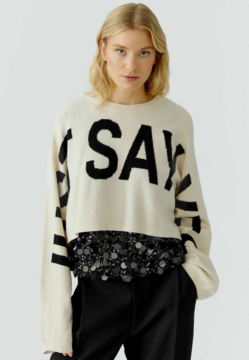 Cropped sweater in off-white with black lettering, long sleeves, and a textured black sequined hem. Soft material and relaxed fit.