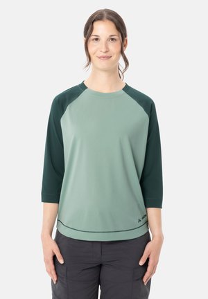 Giovane donna con capelli castani raccolti indossa una maglietta verde a maniche raglan e pantaloni cargo scuri, in piedi con le mani rilassate lungo i fianchi.