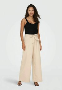 Beige laiad püksid vöödetailiga, kõrge talje ja must tekstureeritud top. Mudel kannab rihmadega musti sandaale. Neutraalne taust.