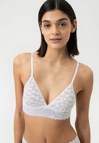 Witte kanten bralette met een driehoekig ontwerp, verstelbare dunne bandjes en een gegolfde kantafwerking aan de onderkant. Gelaagd bloemenpatroon.