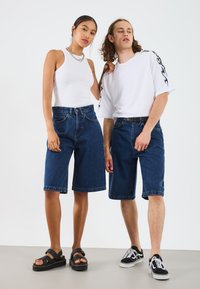 Denim bermudashorts i mörkblå, bar över en vit tank top och en vit t-shirt med svarta flamdetaljer. Svarta sandaler och skor.