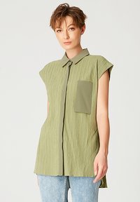 Blouse olive verte à manches courtes en tissu texturé, avec fermeture à boutons, col et poche poitrine unique. Portée avec un jean bleu clair.