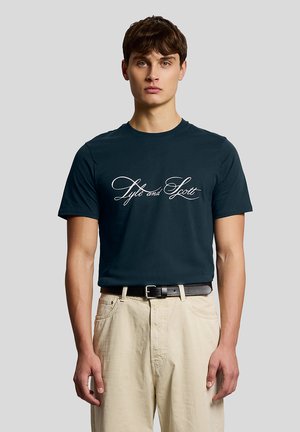 Young man wearing a dark blue T-shirt with "Lyle and Scott" script logo, beige pants, and a black belt, standing against a plain gray background.