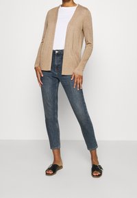 Beige cardigan with long sleeves and open front, paired with blue high-waisted jeans. Black sandals with a silver buckle detail.