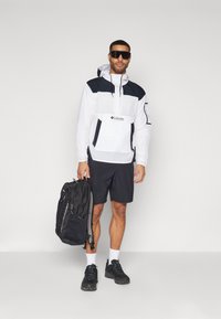 Hvid og sort vindjakke med hætte, lynlåslommer og logo. Sorte shorts og rygsæk, parret med sorte sko og hvide sokker.