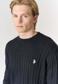 Maglione nero a coste con collo a girocollo a coste, caratterizzato da un piccolo logo bianco sul petto a sinistra. Realizzato in materiale a maglia texturizzata.