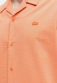 Κοντινή λήψη ροδακινί καρό πουκάμισου με μικρό κεντημένο λογότυπο κροκόδειλου στο στήθος.