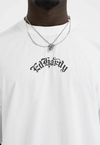 White cotton t-shirt with “Ed Hardy” logo in black font. Features layered silver chain necklaces with a lion skull pendant.