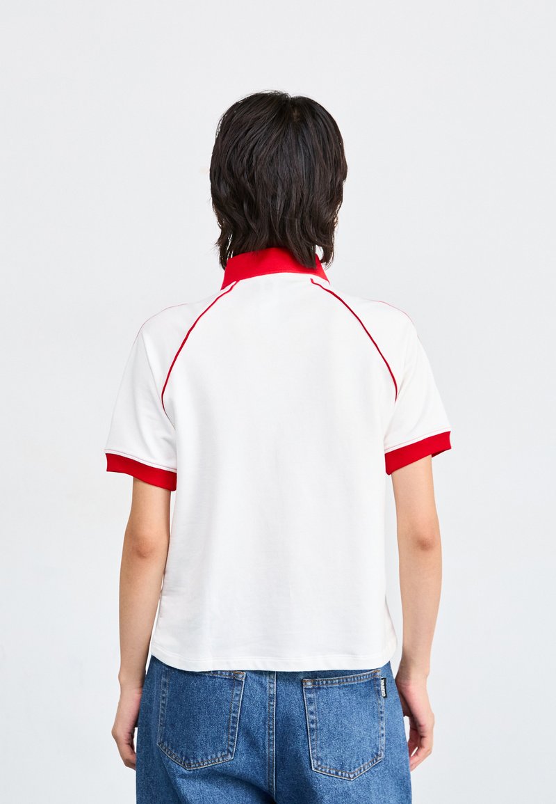 Biała koszulka polo z krótkim rękawem, z czerwonym kołnierzykiem i mankietami, z czerwonymi akcentami w szwie. Krótki fason z gładką teksturą.