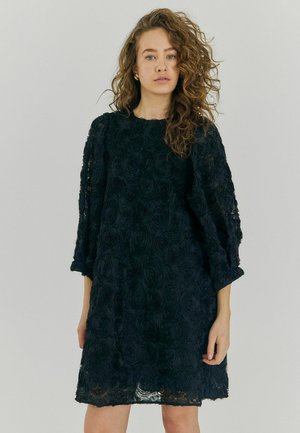 Rochie neagră dintr-un material texturat cu un model de trandafiri. Dispune de mâneci loose și un decolteu rotund, căzând deasupra genunchiului.