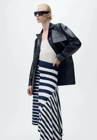 Zwarte leren jas met een glanzende afwerking, gedragen over een wit top en een asymmetrische rok met navy en witte strepen. Zonnebril maakt de look compleet.