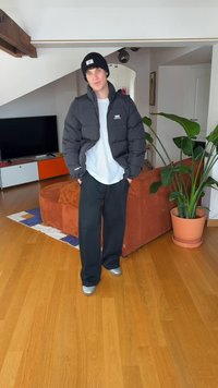 Noor inimene, kes kannab musta mütsi, musta sulejope, valget särki ja musta pükse, seisab elutoas, millel on puidust põrand ja lähedal asuv potitaim.