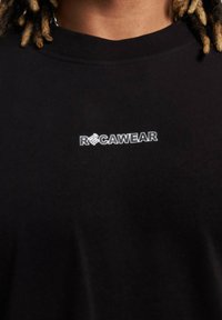 Juodas medvilnės marškinėlis su apvaliu kaklu. Krūtinėje pavaizduotas baltas "RØCawear" logotipas. Paprastas, kasdienis dizainas be raštų.