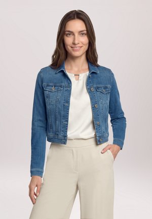 Femme souriante aux longs cheveux bruns porte une veste en jean bleue, un chemisier blanc et un pantalon beige, debout avec une main dans la poche.
