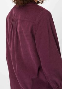 Burgundyröd kordflorsskjorta med ryggsömnad och veck, med långa ärmar och en texturerad yta. Enkel design utan synliga dekorationer.