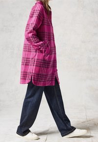 Abrigo de cuadros fucsia en tela suave con mangas largas, bolsillos laterales y dobladillo ligeramente curvado, combinado con pantalones vaqueros oscuros y zapatos blancos.