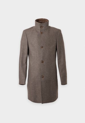 Homme portant un manteau gris, un pull torsadé crème, un jean noir délavé et des mocassins noirs avec des chaussettes blanches, debout les mains dans les poches.