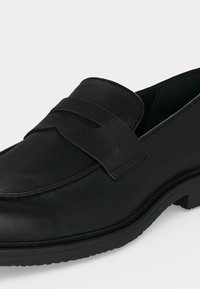 Mocassin en cuir noir à enfiler avec un bout arrondi, un talon bas et une bride décorative sur le dessus.