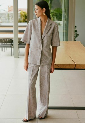 Woman wearing a light gray patterned short-sleeve blazer and matching wide-leg pants, standing indoors beside a wooden table.