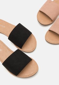 Sorte og beige slip-on sandaler med en bred suedestrop, glat sål og syninger. Den sorte par har en tekstureret overflade.