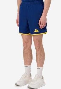 Pantaloni sportivi blu con rifiniture gialle e logo, vita elastica e una superficie liscia. Abbinati a scarpe da ginnastica bianche e calzini.