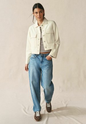 Frau trägt eine cremefarbene Jacke, gestreiftes Hemd, lockere blaue Jeans und braune Turnschuhe, geht mit einer Hand in der Tasche vor einem schlichten Hintergrund.