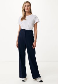 Marineblauwe wijde broek met een zachte textuur, hoge taille en knoopdetail aan de voorkant, gecombineerd met een eenvoudig wit T-shirt en sportieve schoenen.