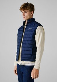 Marine gewatteerd vest met gestikte textuur, hoge kraag en ritssluiting; gedragen over een lichtbeige hemd met knoopmanchetten.