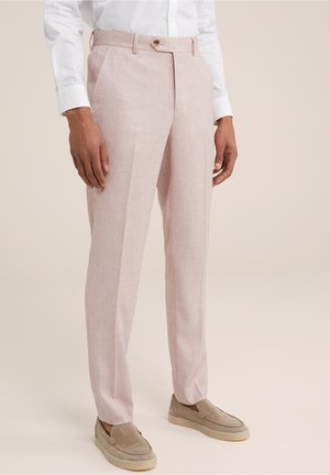 Man wearing light pink tailored trousers, beige slip-on shoes, and a white long-sleeve shirt against a neutral background.