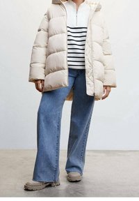 Béžová prošívaná bunda s kapucí, nošená přes bílý a námořnicky pruhovaný svetr, v kombinaci se širokými modrými džínami a béžovými botami.