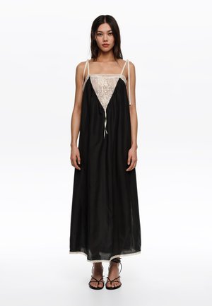 Kobieta z długimi ciemnymi włosami nosi czarną, bezrękawnikową sukienkę maxi z kremową koronkową górą i czarne sandały na paski, stoi na tle białego tła.