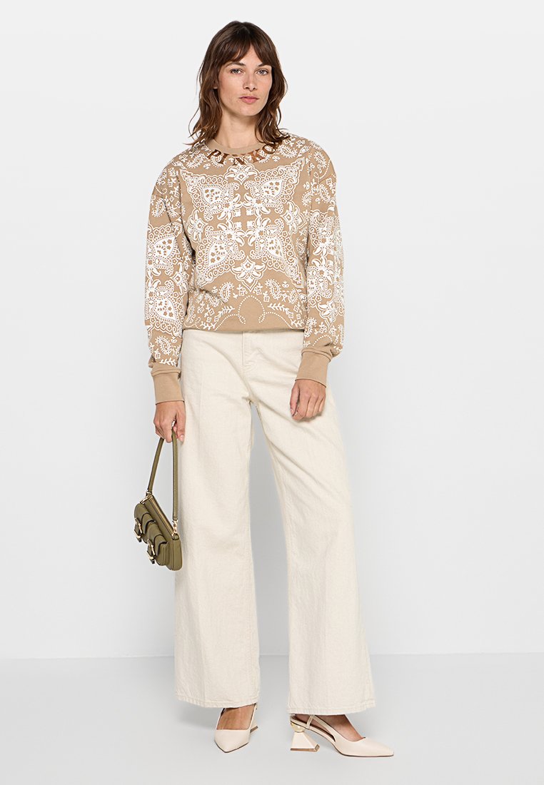 Beige sweatshirt met een wit bloem- en paisleypatroon, ribgebreide manchetten, gecombineerd met crèmekleurige broek met wijde pijpen en beige hakken, terwijl een groene tas wordt vastgehouden.