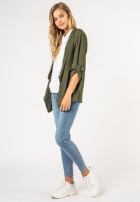Olijfgroene open-front jas met opgerolde mouwen, gecombineerd met een wit T-shirt en lichtblauwe skinny jeans. Witte sneakers maken de look af.