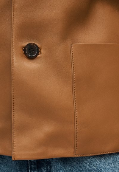 Indumento in pelle color cuoio con un bottone marrone scuro e una tasca cucita sopra il tessuto in denim blu.