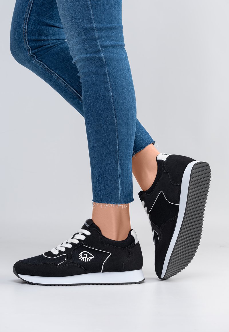Benen met cropped blauwe spijkerbroeken en zwarte sneakers met witte veters en zolen, één voet plat op de grond en één opgetild, waarbij het loopvlak van de schoen zichtbaar is.