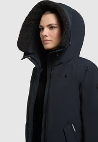 Marine vinterjakke med en stor, fuzzy-foret hætte, lynlås lukning, sidelommer og et broderet bladlogo på brystet. Blød tekstur.