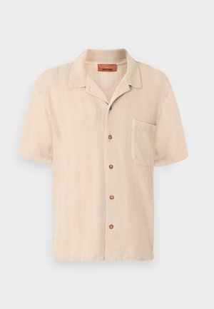 Bēša īss piedurkņu krekls ar pogām, teksturētu zivssānu auduma rakstu, priekšējo krūšutvertnes kabatu un mīkstu apkakli.