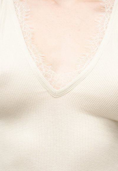 Κλείσιμο εικόνας ριμπ μπεζ ρούχου σε σχήμα V με λεπτή δαντέλα στο ντεκολτέ, που δείχνει μέρος του άνω στήθους ενός ατόμου.