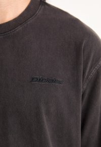 Temno siva majica s krojem po vratu, izdelana iz bombaža, z teksturiranim črnim vezenim logotipom "Dickies" na prednjem delu in sproščenim krojem.