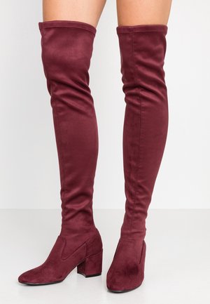 Lårhøje, mørkerøde støvler i ruskind med blokhæl båret på bare ben mod en enkel lys baggrund.